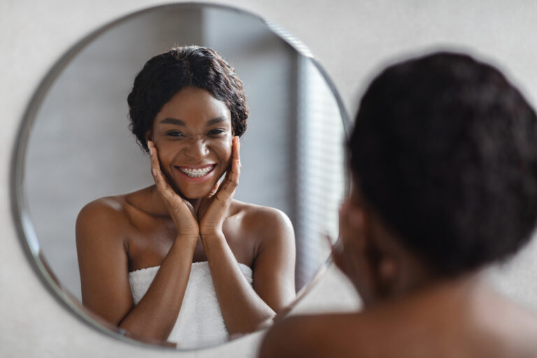 A black woman smiles at herself in a mirror noticing HydraFacial benefits in Kansas City