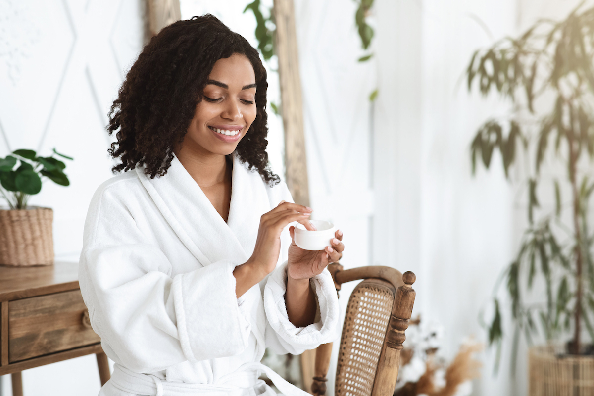 A woman in a white robe dips her finger into a tub of skincare product
