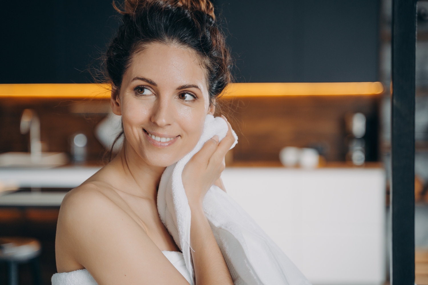 A smiling woman wearing a towel wipes her face off with another white towel while looking off to the side