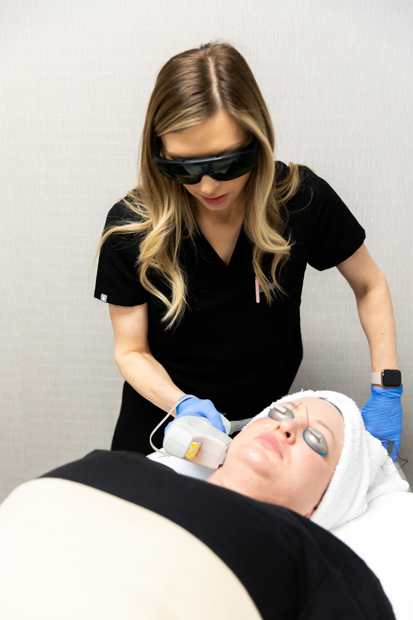 Female client is receiving facial treatment from Kansas City Skin and Cancer Center professional. Her face is covered in thin layer of clear gel and she is wearing protective eye coverings. Aesthetician is visible in this shot.