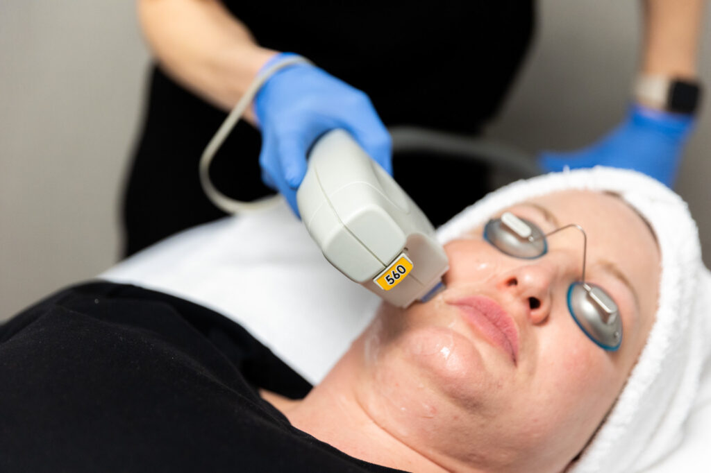 Female client is receiving facial treatment from Kansas City Skin and Cancer Center professional. Her face is covered in thin layer of clear gel and she is wearing protective eye coverings. Aesthetician is visible in this shot.