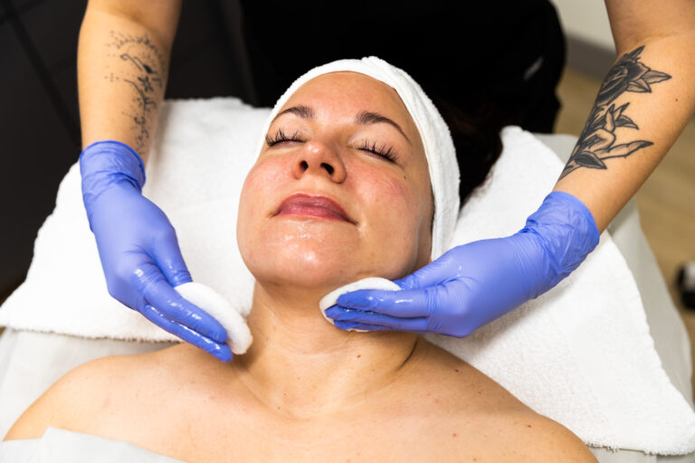 A Kansas City Skin and Cancer Center employee uses two cotton pads on a client's face during the facial treatment. The client is lying down with her eyes closed.