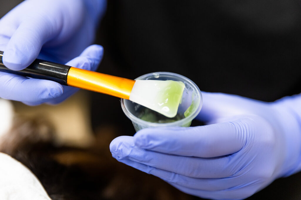 A pair of gloved hands holds a small cup of facial application. The gel is green, and one of the hands is holding a smooth applicator.