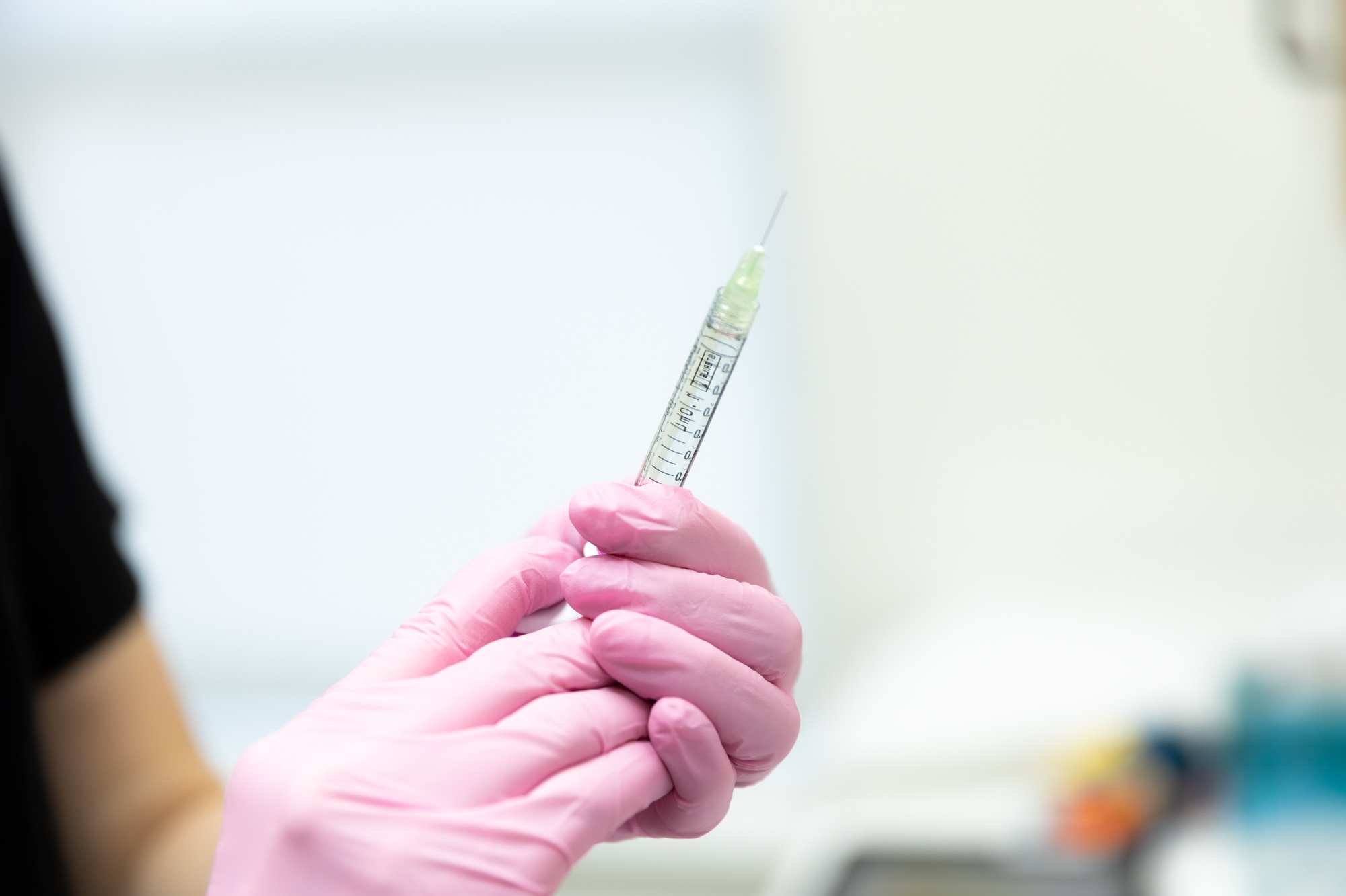Image is of a pair of gloved hands holding up a syringe. The needle sticks out of the top of the syringe.