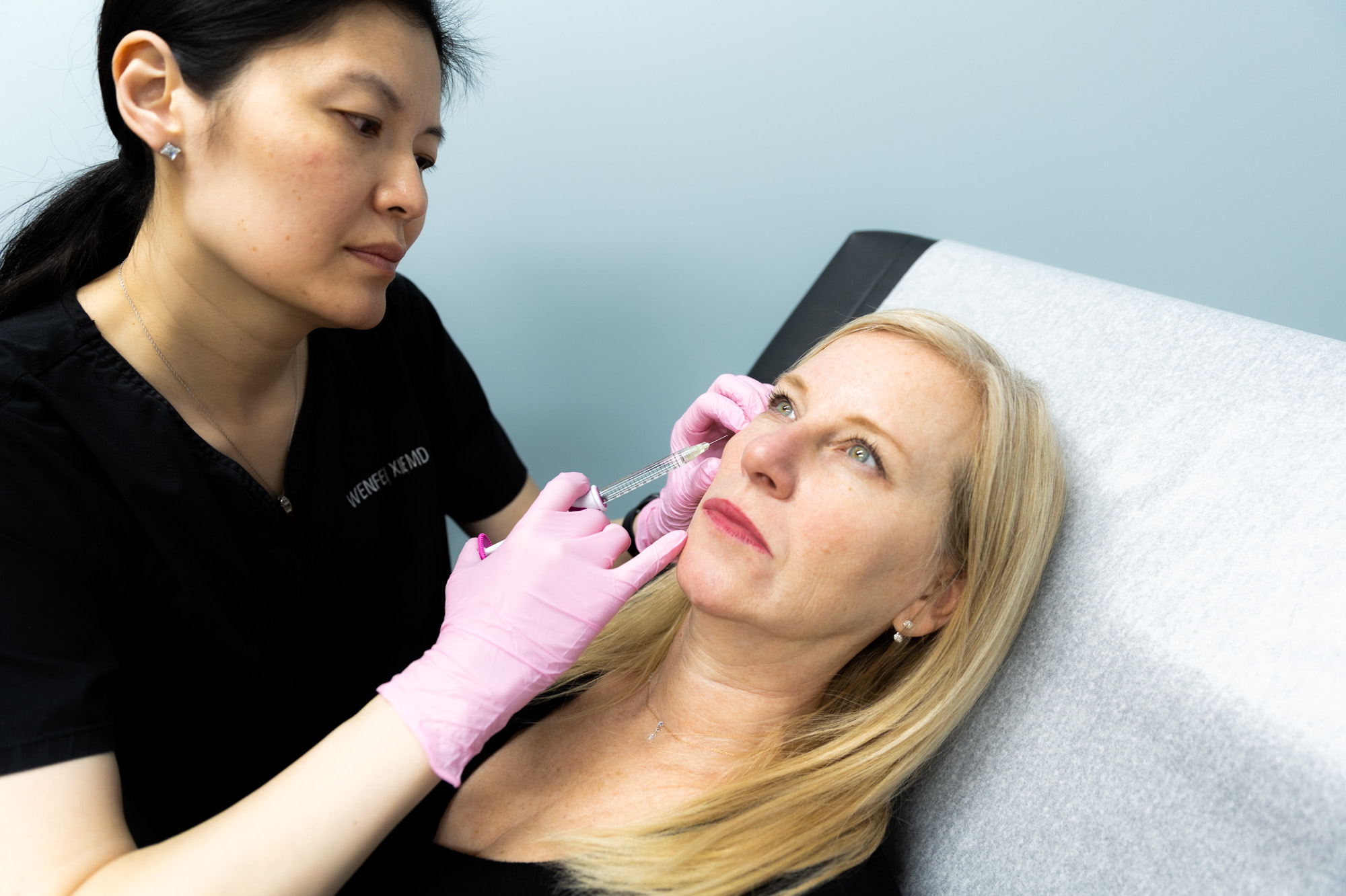 A Kansas City Skin and Cancer Center doctor injects a filler into a patient's right cheek, below her eye. The doctor uses a syringe with a needle.