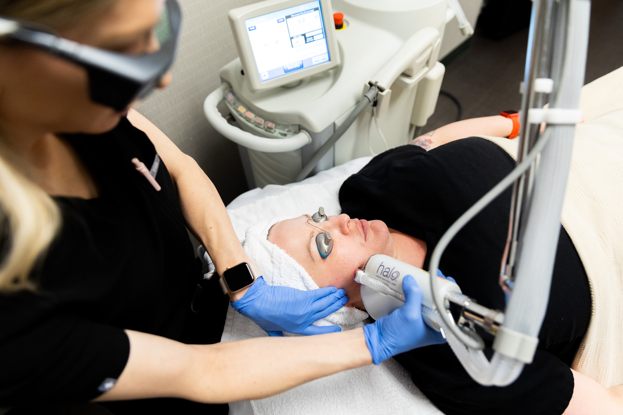 A Kansas City Skin and Cancer Center aesthetician holds the HALO treatment device against the female client's right jawline. The client is lying down with protective eye coverings on.