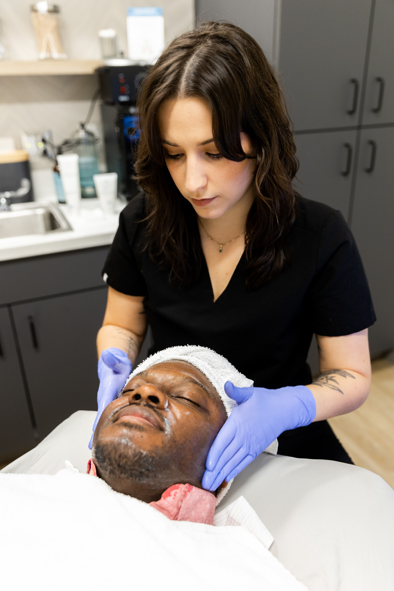 A female medical provider gently holds a male client's head in her hands as she finishes his hydrafacial treatment. The client is lying down with his eyes closed.