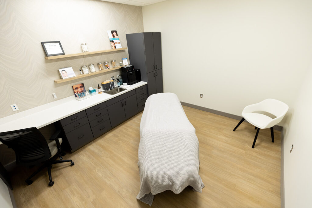 An examination and treatment room. There is an examination chair at the center of the room, covered by a blanket. There is an extra chair in the back right corner. The left side of the room has cabinets and countertops with a variety of different bottles and flyers.