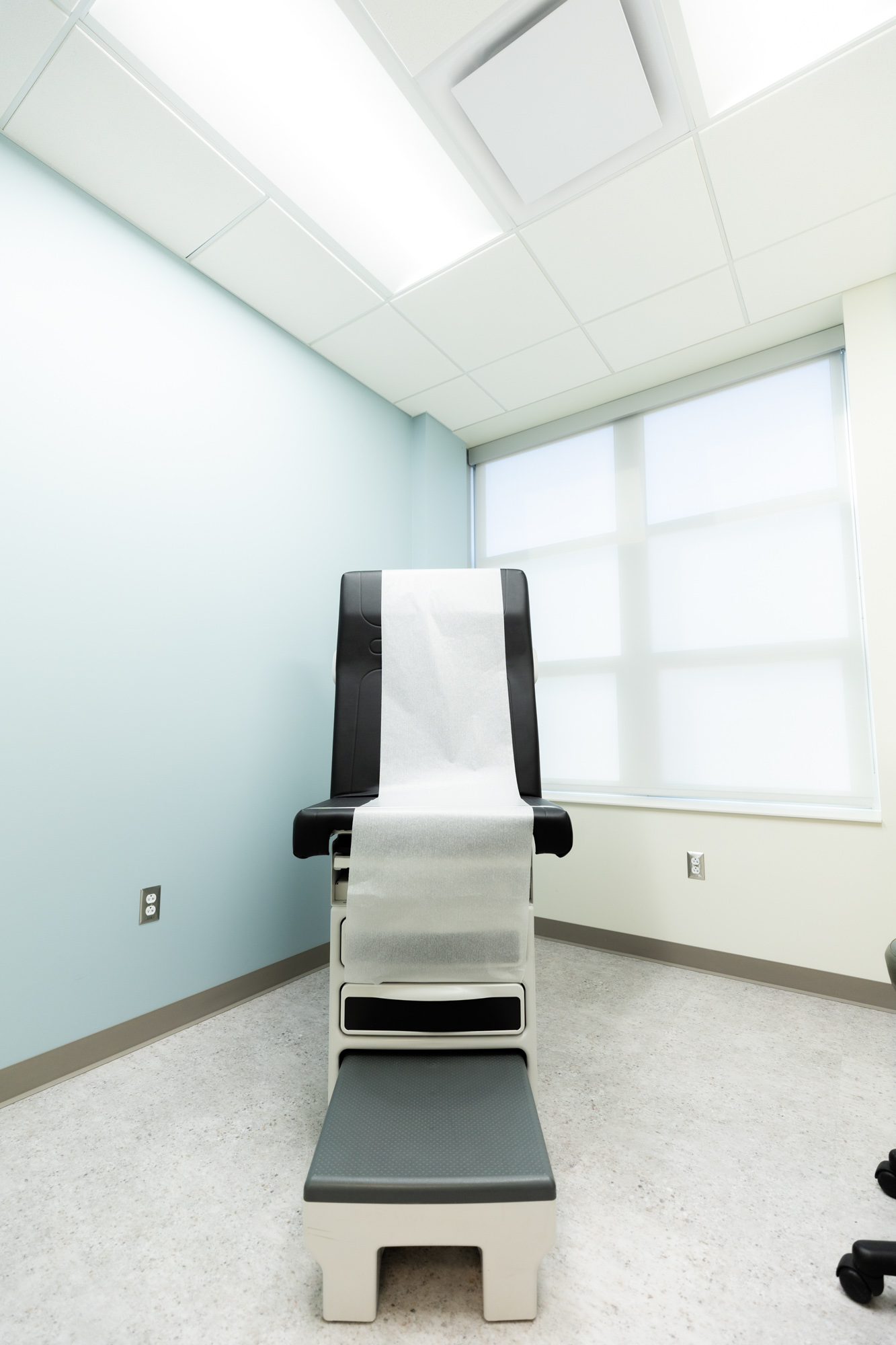 Image shows one corner of an examination and treatment room. There is a medical examination chair with a layer of thin white paper over it, much like a chair you might see at a regular physician's office. There is a window in the back but the blinds are pulled down.