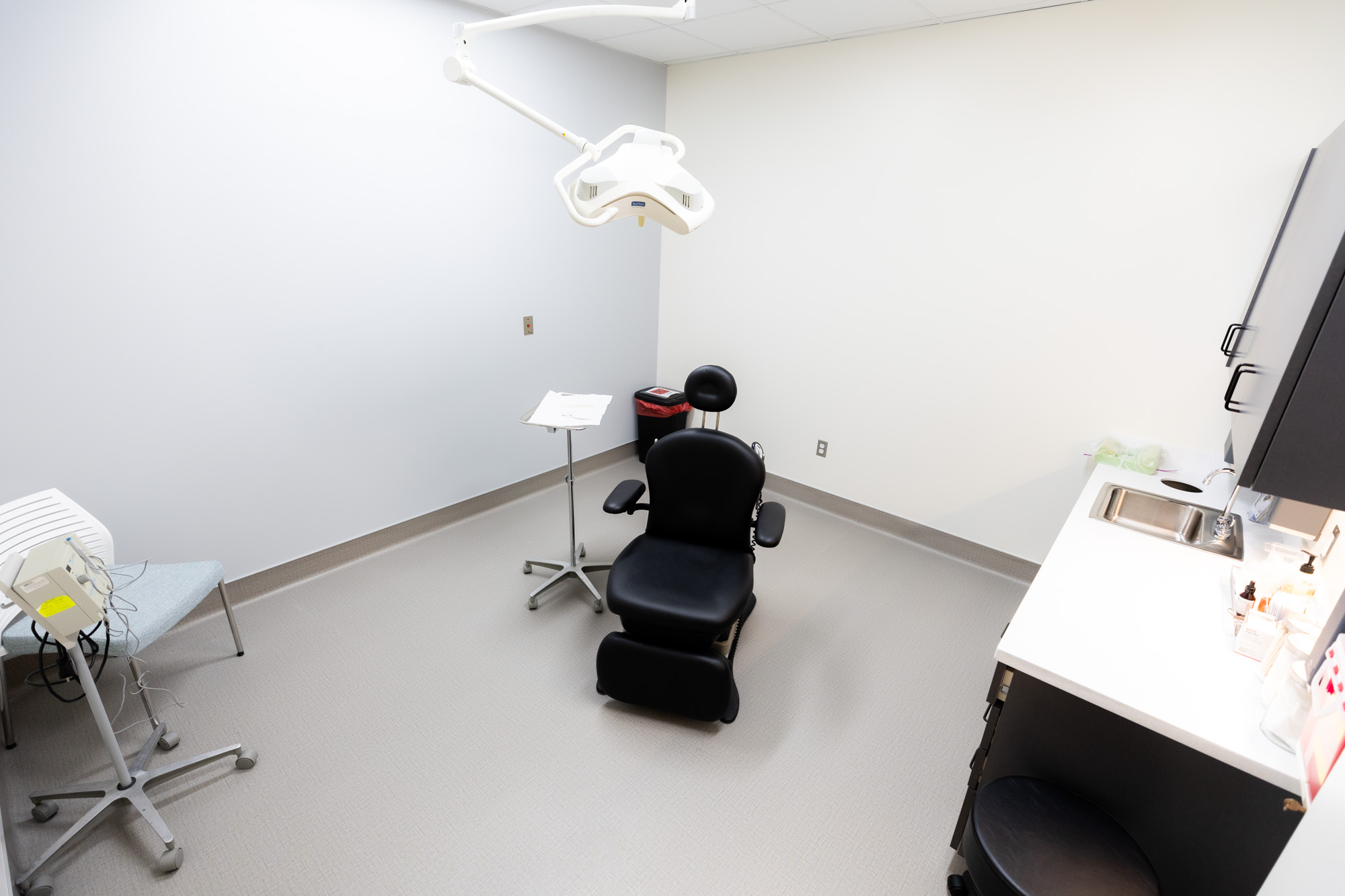 Image of a treatment room. In the middle of the room is a sleek black medical examination chair with a rolling side table next to it. There is also an overhead lighting device, similar to what is at a dentist's office. In one corner is an extra chair and then a rolling machine with several wires coming out of it. On the other wall is a countertop with a sink and cabinets up above it.