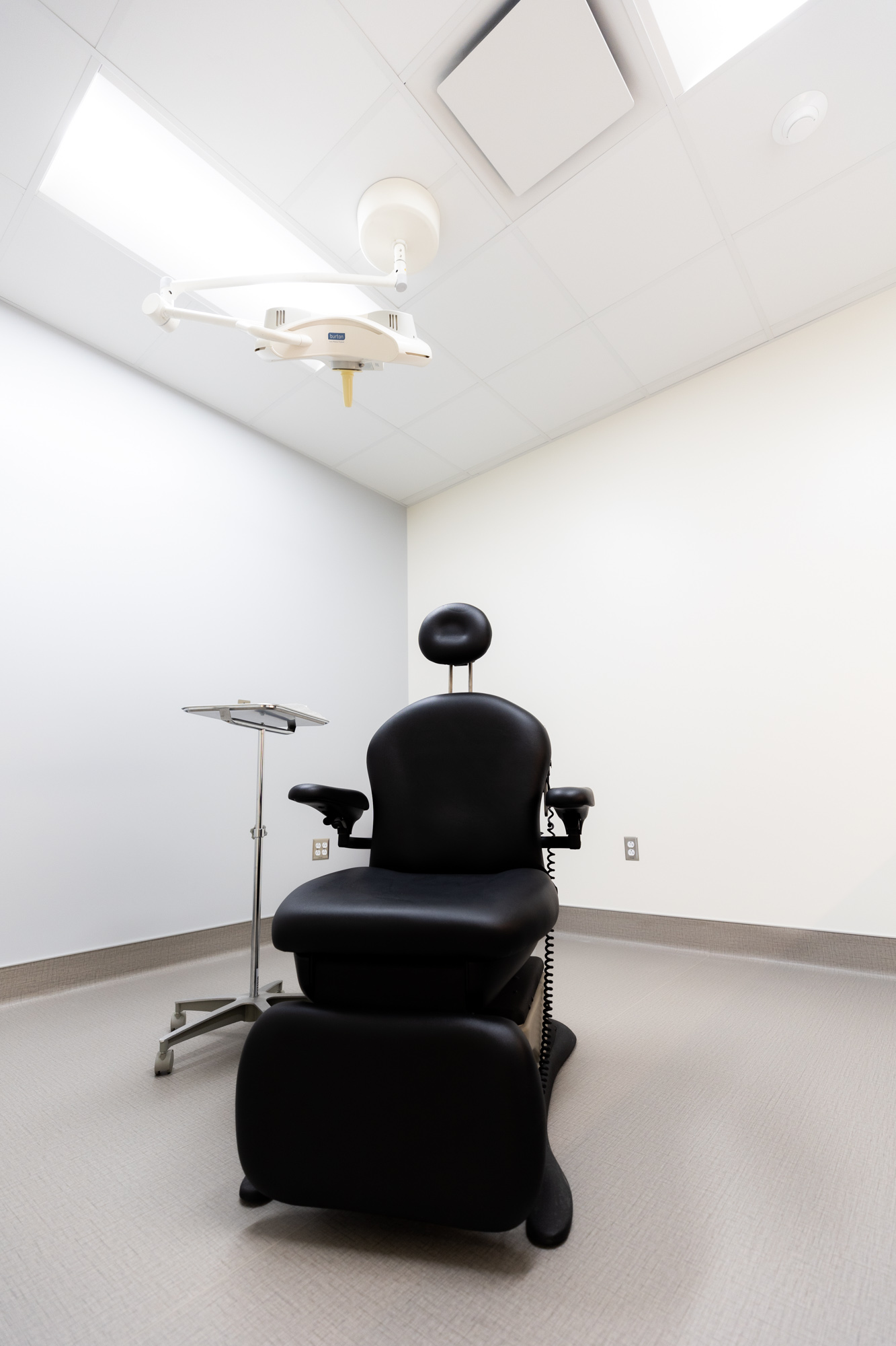 Image of a treatment room. In the middle of the room is a sleek black medical examination chair with a rolling side table next to it. There is also an overhead lighting device, similar to what is at a dentist's office. Only the middle and one corner of the room are visible.