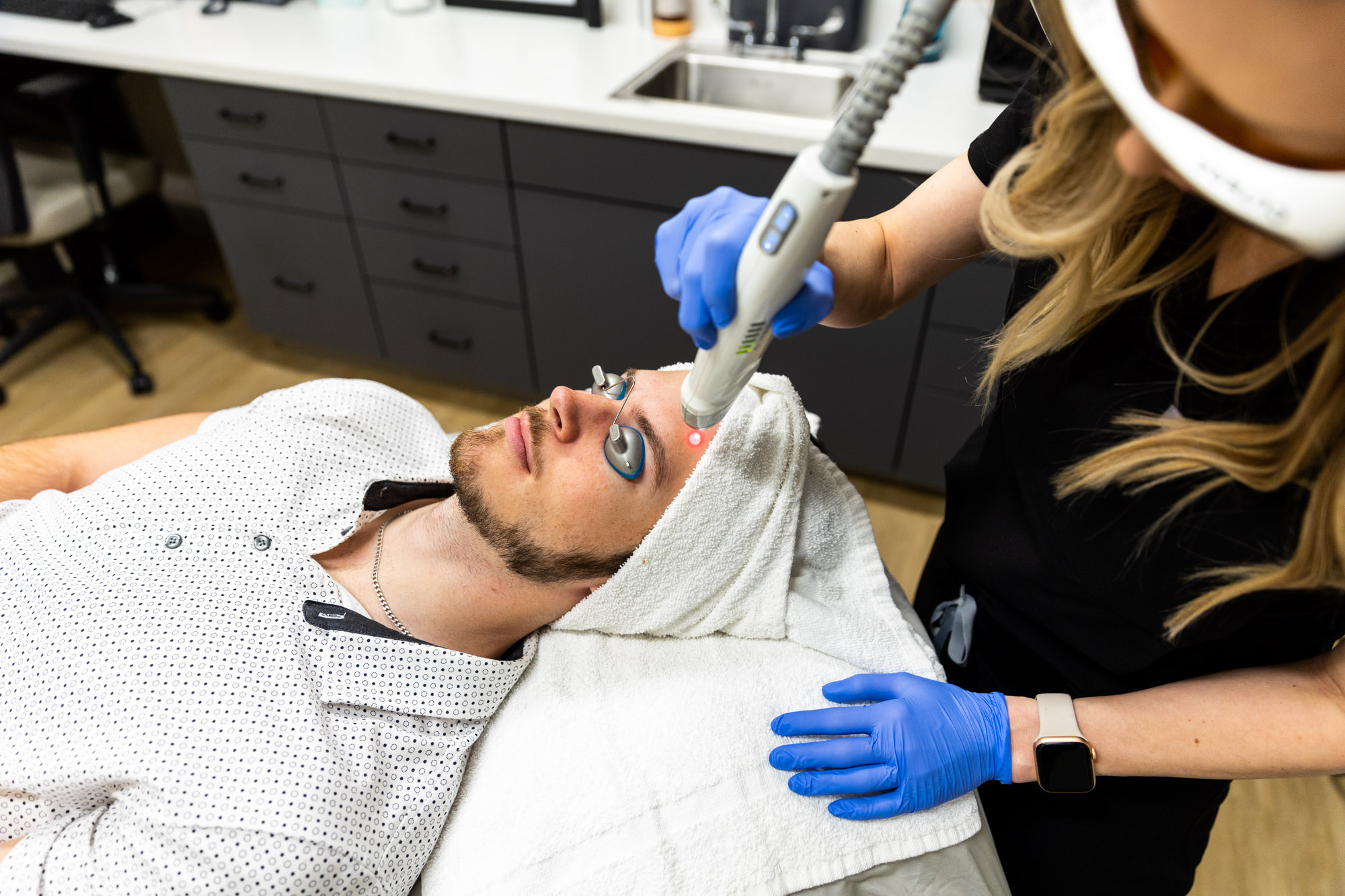 A female medical provider holds the Laser Genesis tool over a male client's forehead. He's wearing protective eye coverings and a towel over his hair. The medical provider is wearing protective sunglasses. The image is taken from above.
