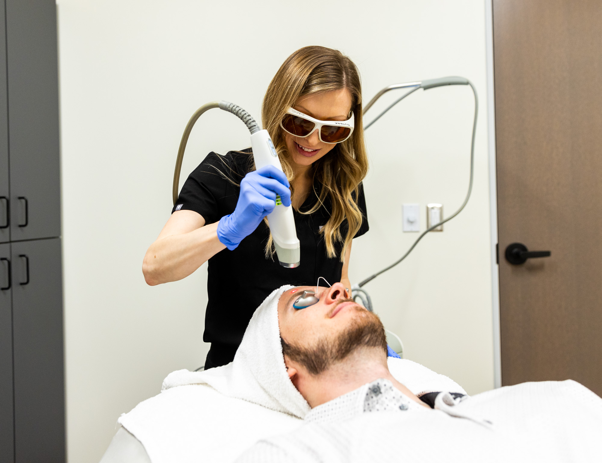 A female medical provider holds the Laser Genesis tool over a male client's forehead. He's wearing protective eye coverings and a towel over his hair. The medical provider is wearing protective sunglasses.