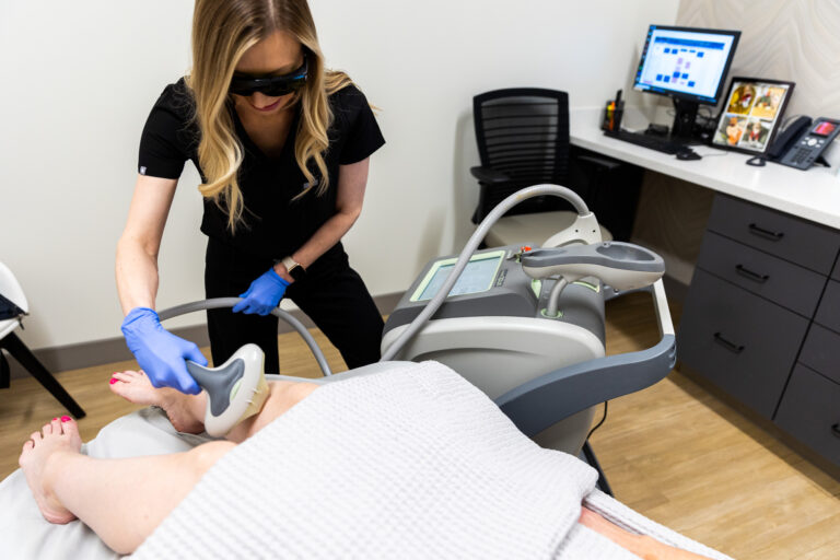 A female aesthetician uses the handheld laser hair reduction device on a client's right leg. Only the client's legs are visible. They are lying down while the treatment for laser hair removal in Shawnee is administered.