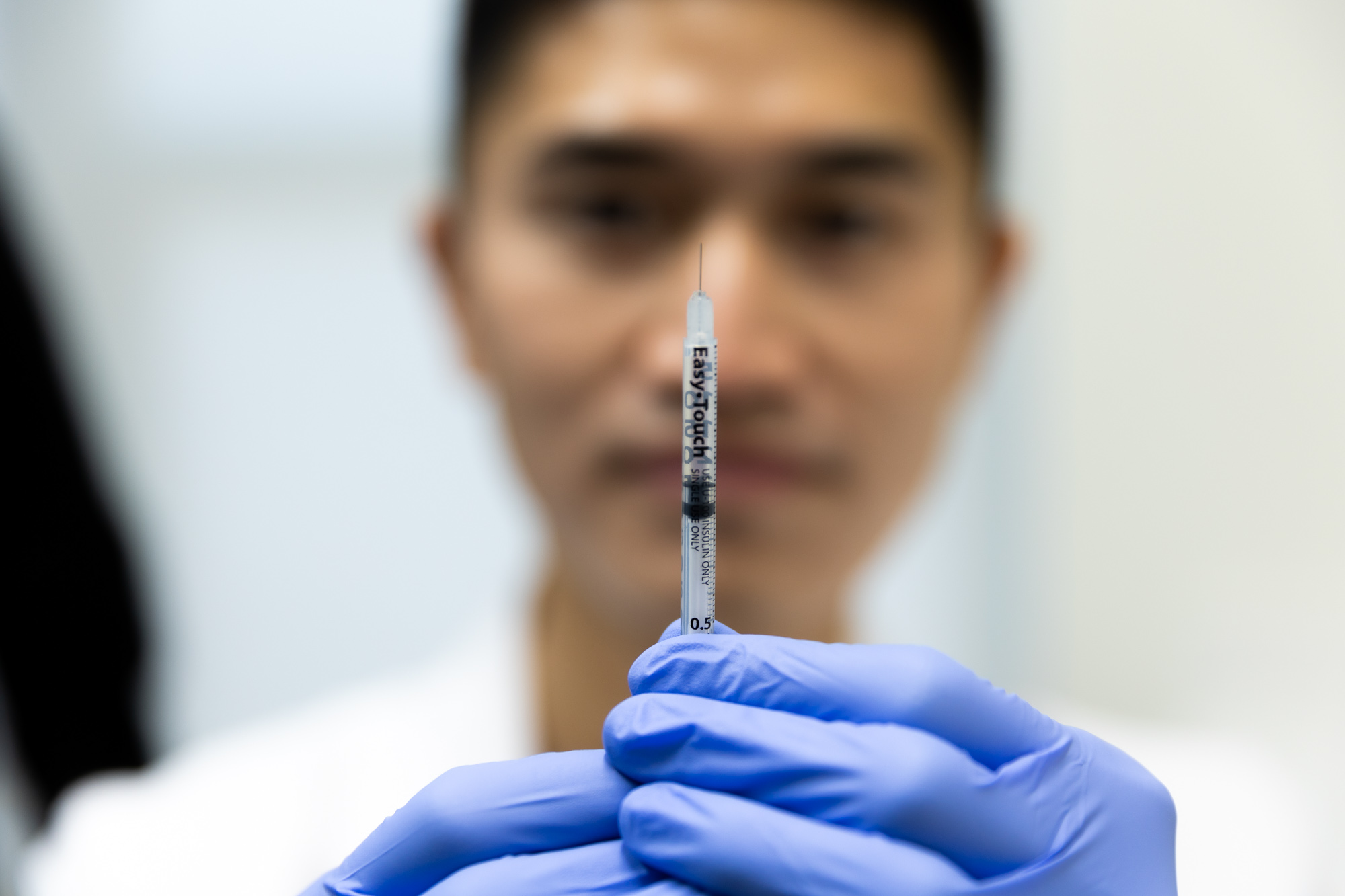 A doctor holds up a syringe with a needle. Syringe reads Easy Touch. The doctor's face is out of focus in the background; he is measuring the contents of the syringe.
