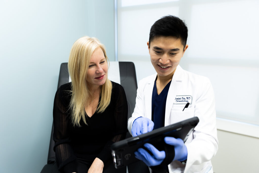 A Kansas City Skin and Cancer Center doctor holds an iPad up and points at something on screen to show to female patient seated beside him.
