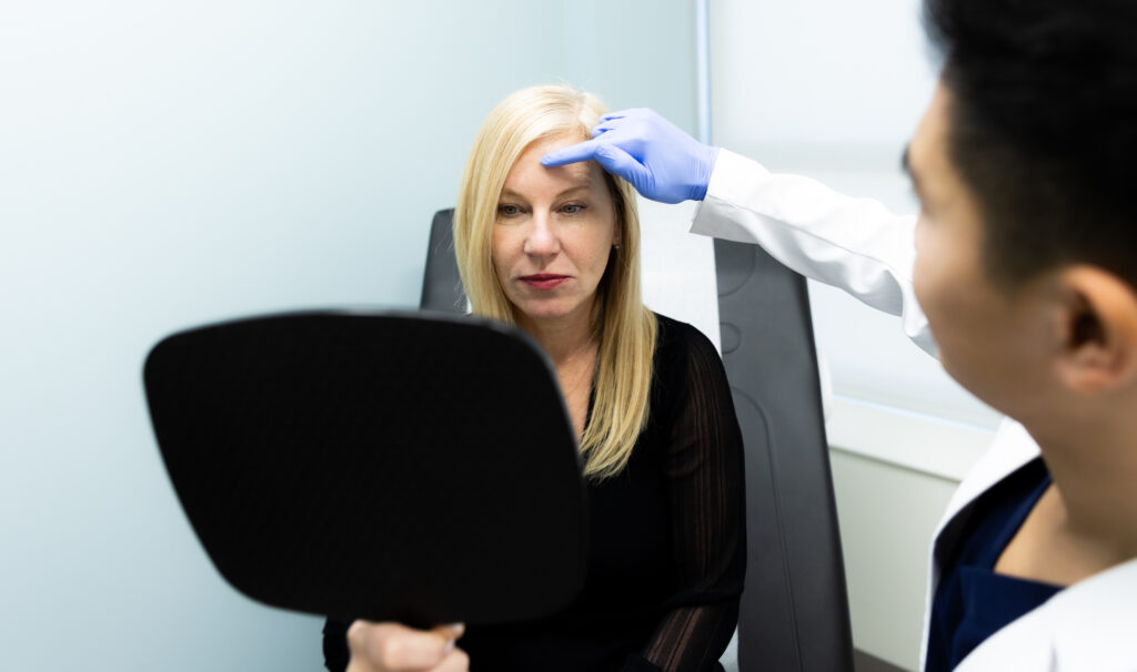 A provider at Kansas City Skin & Cancer Center meets with a patient for her consultation on skin toning in Kansas City, MO. The patient holds a hand mirror as her provider points to areas of discoloration on her forehead.