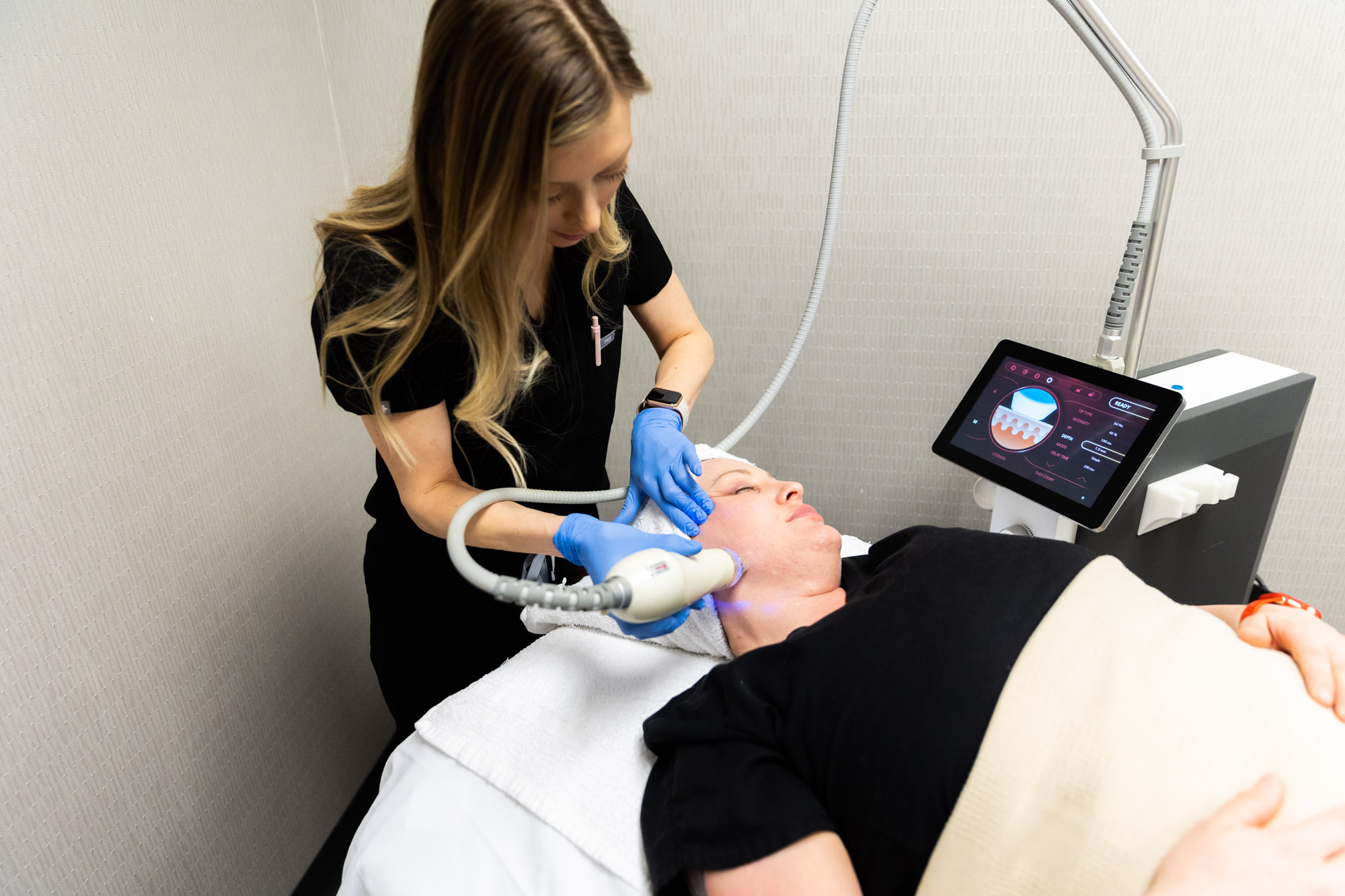 A female client is receiving a microneedling treatment. She is lying on a medical bed with hair covered by towel. Eyes are closed. A female Kansas City Skin and Cancer Center employee is holding the microneedling tool in gloved hands. The machine shows the skin layers.