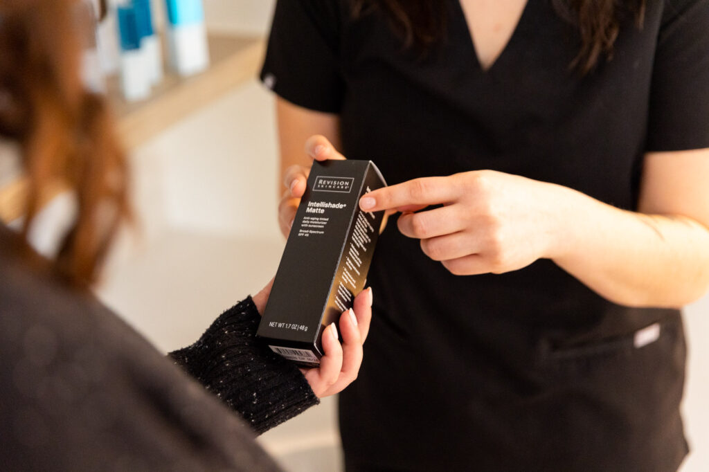 A provider at KCS&CC shows a patient a medical-grade skincare product, usually recommended alongside a rosacea treatment in Kansas City. The black box of the product is visible, with the provider explaining how it can soothe rosacea symptoms.