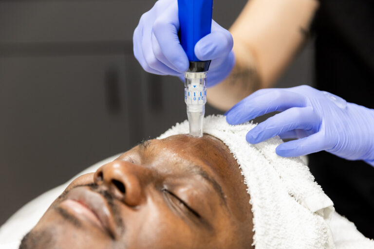 A female medical provider gently holds a male client's head in her hands as she finishes his hydrafacial treatment. The client is lying down with his eyes closed.