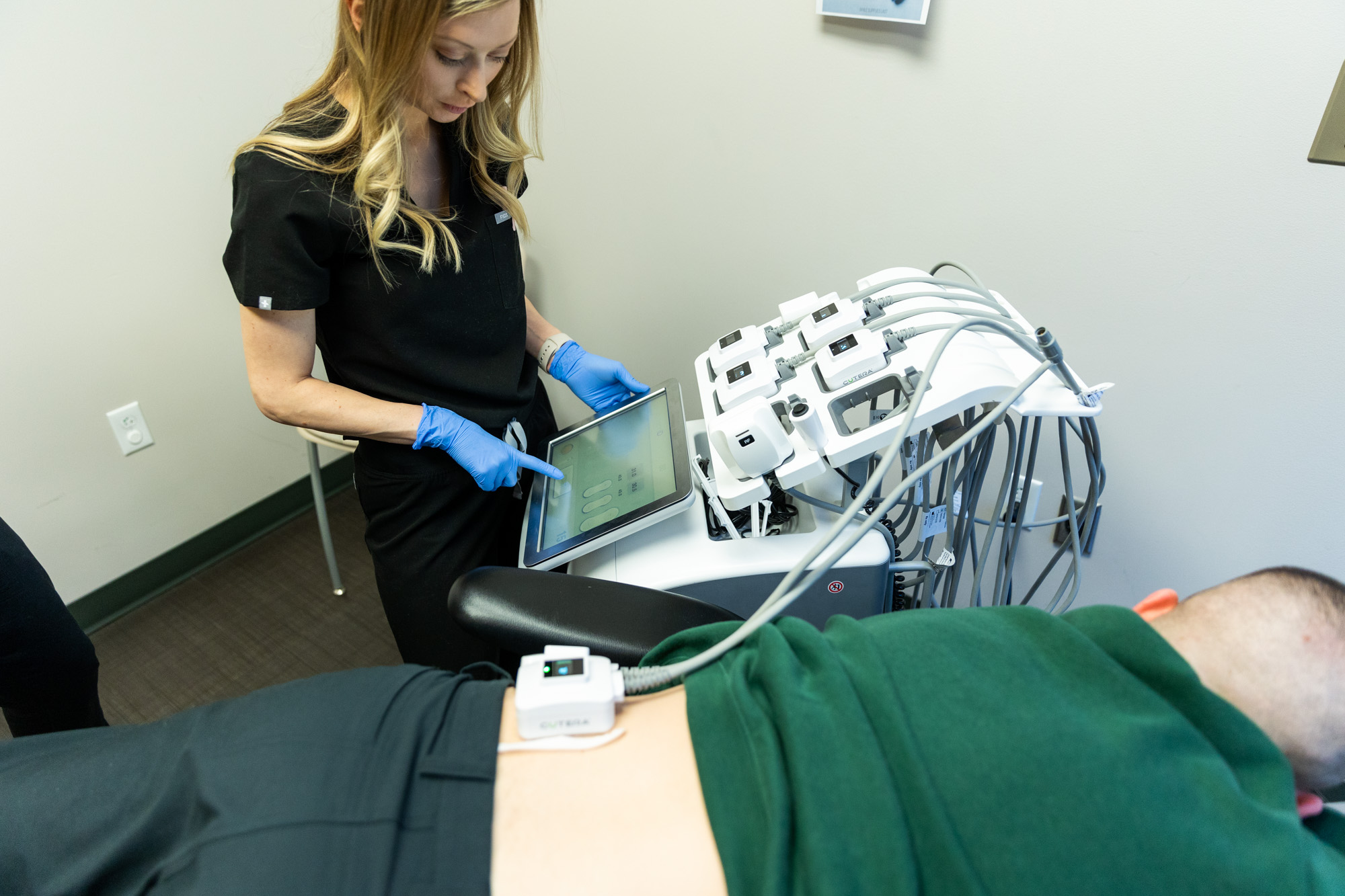 A Kansas City Skin and Cancer Center professional selects a setting on the TruSculpt machine. A male patient is lying down with the Cutera applicator on their lower back.