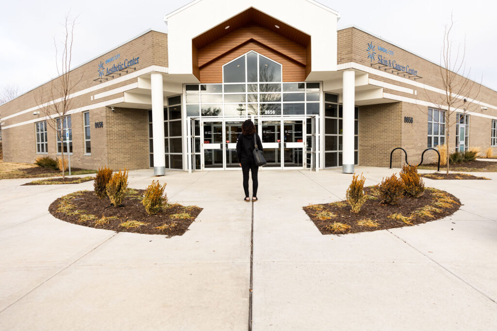 A female client stands outside the front doors of the Kansas City Skin and Cancer Center. One side of the building is labeled Aesthetic Center, the other is labeled Skin and Care Center.