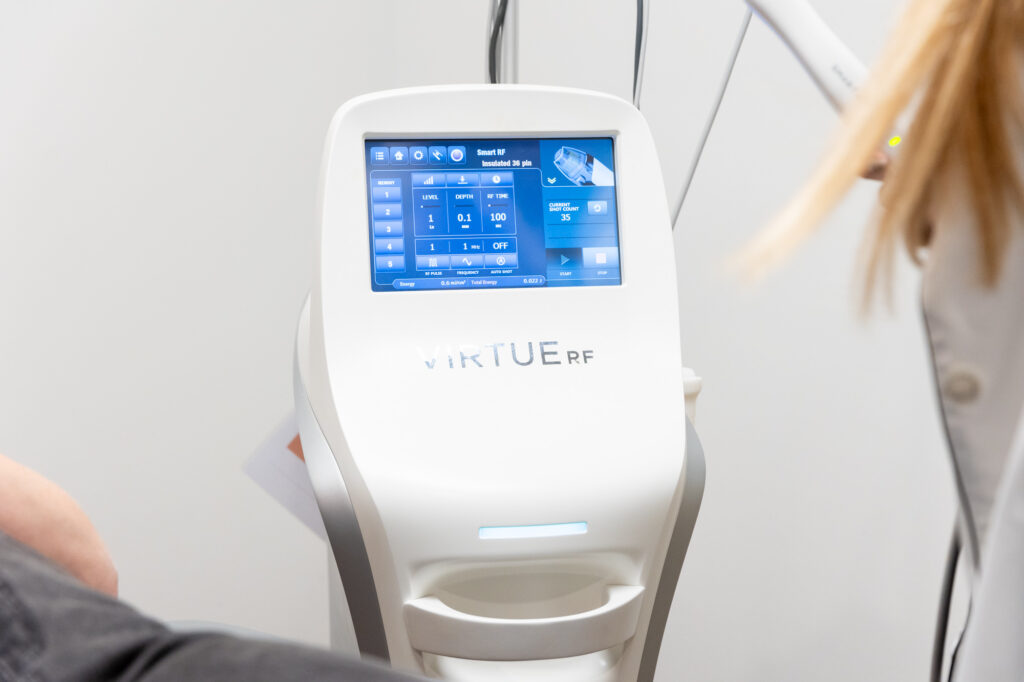 A female client is receiving a microneedling treatment. She is lying on a medical bed with hair covered by towel. Eyes are closed. A female Kansas City Skin and Cancer Center employee is holding the microneedling tool in gloved hands. The machine shows the skin layers.